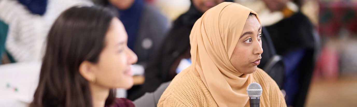 Photograph of a woman holding a microphone and speaking at the Flourishing Communities Learning and Celebration Event. There is a woman sitting besode her that ids out of focus. Photograph by Photographer London: https://freelanceeventsphotographer.co.uk/