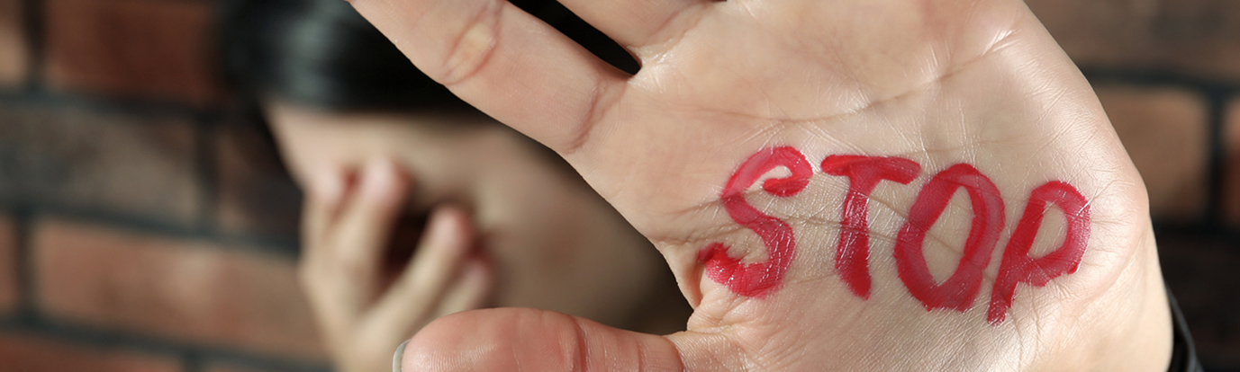 A person holds up a hand toward the camera with 'STOP' written on the palm in red, while in the blurred background another person crouches against a brick wall with their head down, shielding their face.