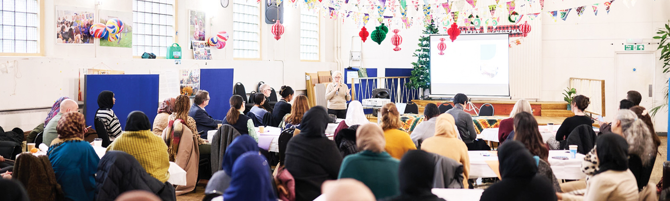 Alison Robert, THCVS Partnerships Manager speaking at the Flourishing Communities Learning and Celebration Event. There is a large seated audience in a community hall decorated with colourful bunting and hanging ornaments, with a presentation slide displayed on a screen behind them. Photograph by Photographer London: https://freelanceeventsphotographer.co.uk/
