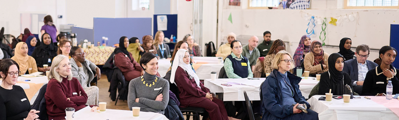 A diverse group of adults seated around tables in a bright community room, listening to a presentation at teh Flourishing Communities celebration event. Photograph by: Photographer London - https://freelanceeventsphotographer.co.uk/coroporate-event-photographer-london.
